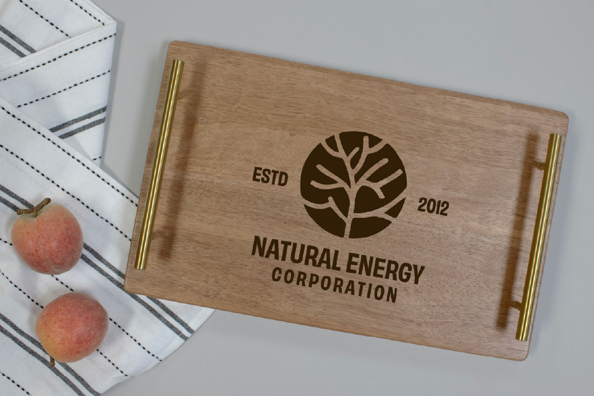 A rectangular wooden serving tray with gold metal handles, featuring an engraved tree logo and the text 'ESTD 2012' and 'NATURAL ENERGY CORPORATION'. The tray is styled on a light gray surface next to a striped white napkin and two peaches.