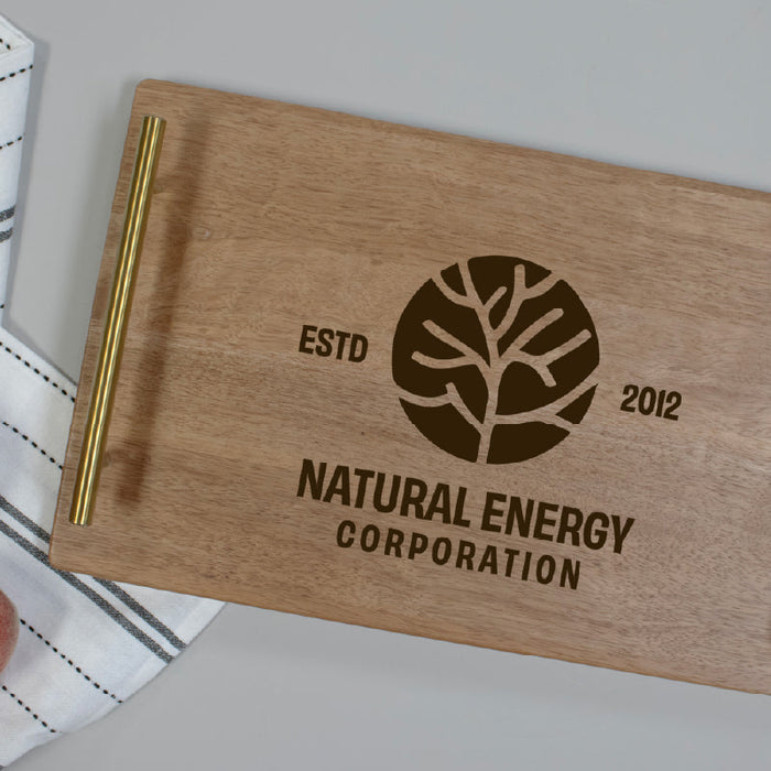 A rectangular wooden serving tray with gold metal handles, featuring an engraved tree logo and the text 'ESTD 2012' and 'NATURAL ENERGY CORPORATION'. The tray is styled on a light gray surface next to a striped white napkin and two peaches.
