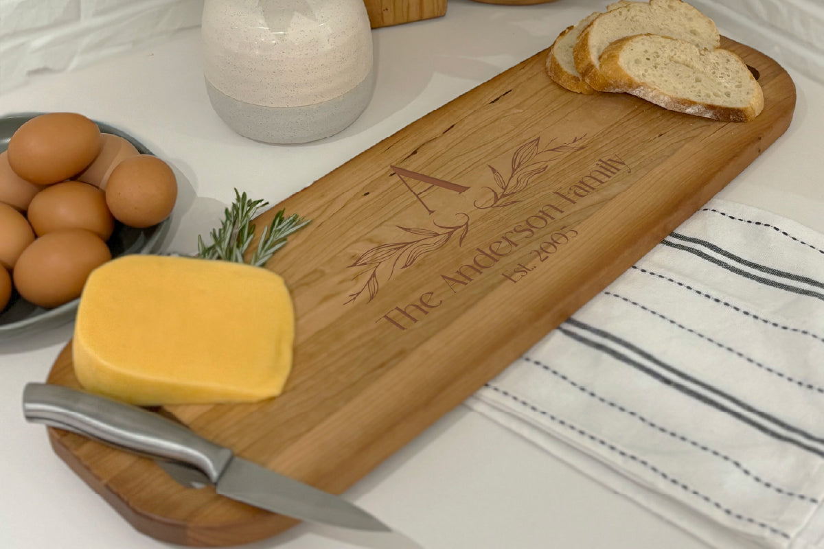 A personalized cutting board sitting on a white table with bread and cheese on it. The board is engraved with a family name, established date, and a monogram.