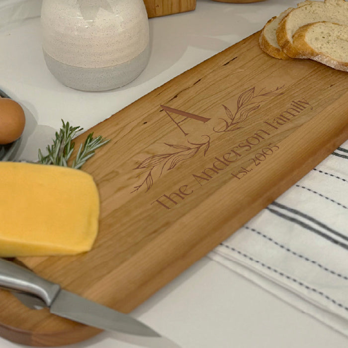 A personalized cutting board sitting on a white table with bread and cheese on it. The board is engraved with a family name, established date, and a monogram.