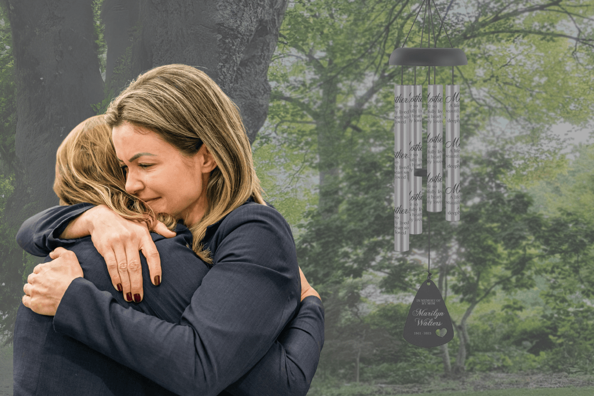 Two women hug during time of grief next to memorial wind chime for mother