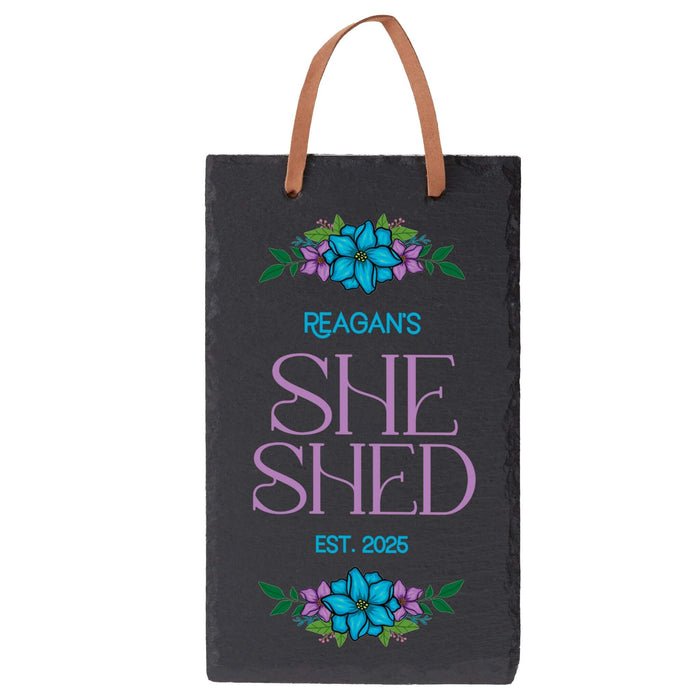 Slate sign with floral designs and text 'Reagan's SHE SHED EST. 2025' on a white background.