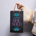 Person holding a personalized 'She Shed' sign with floral designs on a plain background