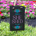Personalized 'SHE SHED' sign with floral designs in a garden setting