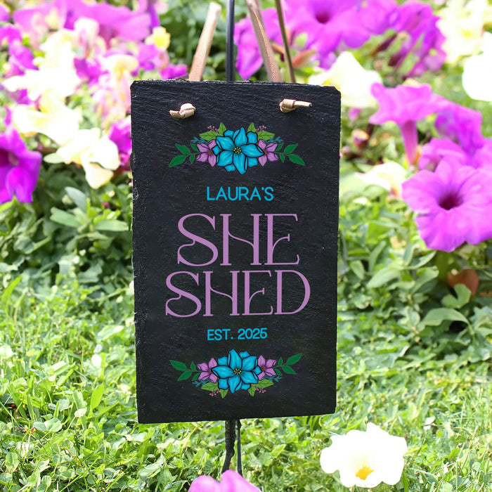 Decorative sign with 'Laura's She Shed' text in a garden setting with pink flowers