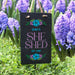Decorative sign with 'EMILY'S SHE SHED EST. 2025' in a garden setting with purple flowers.