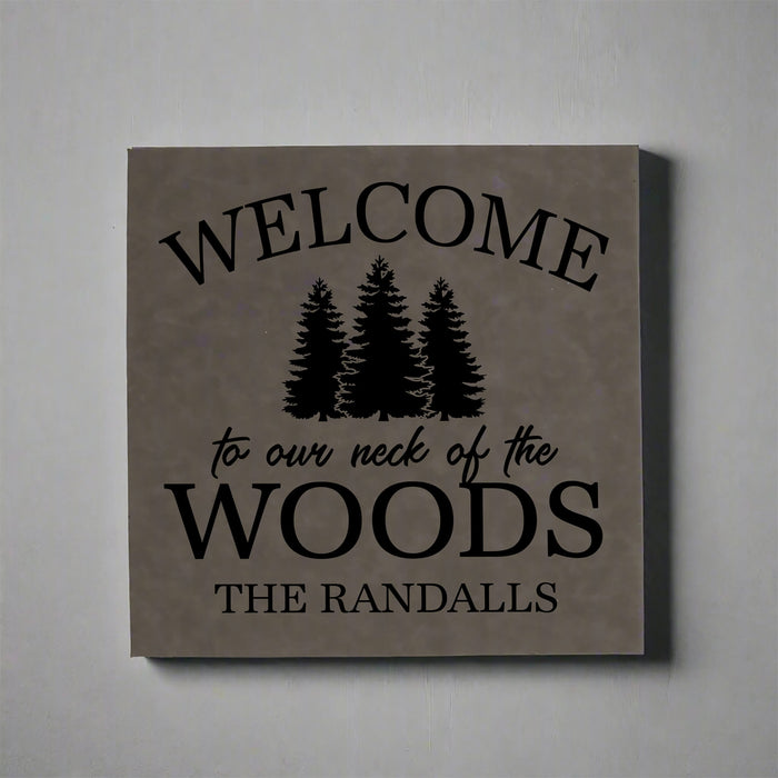 gray vegan leather wall sign engraved  with 'Welcome to our neck of the woods' text and tree graphics on a gray background.