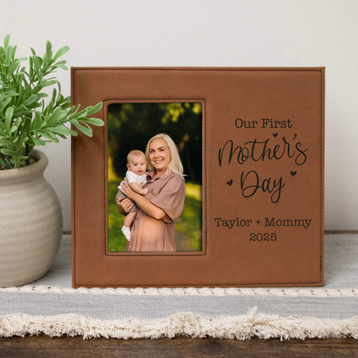 Brown leather photo frame with a picture of a woman holding a baby, text 'Our First Mother's Day Taylor + Mommy 2025', on a wooden surface with a plant.