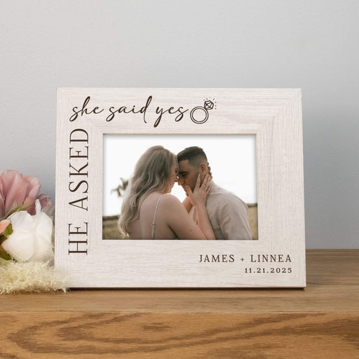 Personalized photo frame with a couple embracing and text 'She asked, He said yes' on a wooden surface.