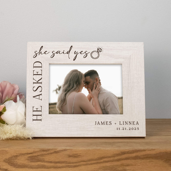 Personalized photo frame with a couple embracing and text 'She asked, He said yes' on a wooden surface.
