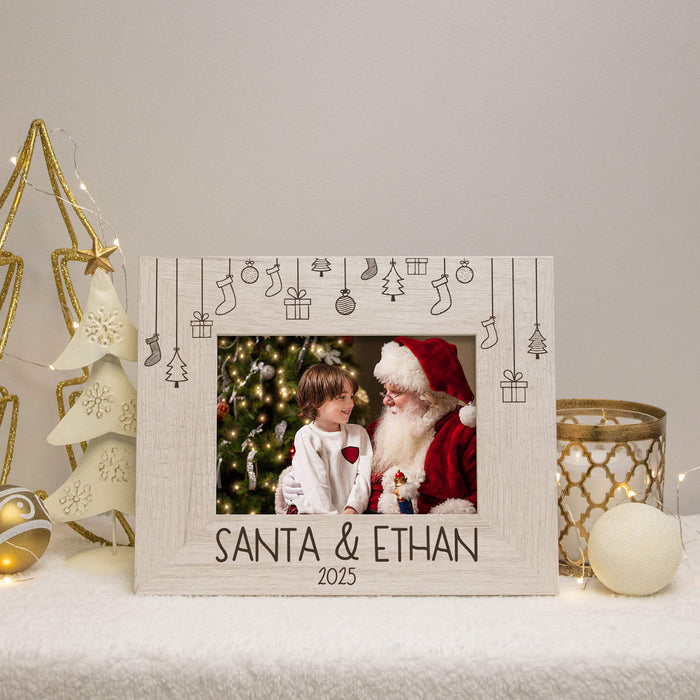 A personalized Christmas photo frame with a rustic white wood finish, featuring engraved holiday icons and the text 'SANTA & ETHAN 2025' at the bottom. The frame is displayed on a white surface with festive holiday decorations, including a gold star tree, a ceramic Christmas tree, and a gold patterned candle holder.