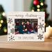 A personalized Christmas photo frame with a distressed white wood finish, featuring engraved brown snowflakes, the text 'merry christmas' at the top, and 'THE STAPLETONS' at the bottom. The frame is shown on a wooden table with a blurred festive background of a Christmas tree and candles.