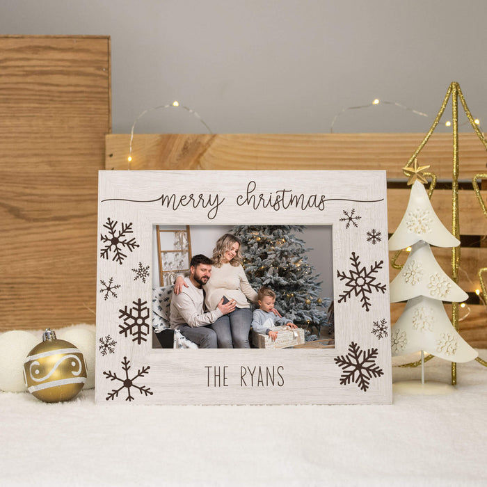A personalized white wooden Christmas picture frame with 'merry christmas' and 'THE RYANS' engraved on it, featuring laser-engraved snowflakes. The frame is styled on a white surface with a gold ornament, a wooden crate, and a white tree light in the background.