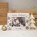 A personalized white wooden Christmas picture frame with 'merry christmas' and 'THE RYANS' engraved on it, featuring laser-engraved snowflakes. The frame is styled on a white surface with a gold ornament, a wooden crate, and a white tree light in the background.