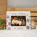 A personalized Christmas photo frame with a distressed white wood finish, decorated with brown snowflakes and the text 'merry christmas' and 'THE CAMERONS'. The frame is shown standing on a white faux-fur surface, flanked by a pine branch with red berries on the left and a lit gold-patterned candle holder on the right, with a wooden slat wall and fairy lights in the background.