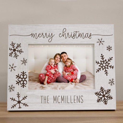 A personalized Christmas photo frame with a distressed white wood finish, featuring 'merry christmas' at the top, snowflake designs on the sides, and the family name 'THE MCMILLENS' engraved at the bottom. The frame is shown holding a photo of a family of four on a bed.