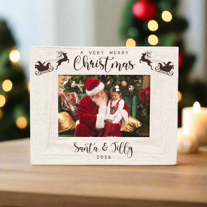 A personalized white wooden Christmas photo frame on a wooden table, featuring laser-engraved text reading 'A VERY MERRY Christmas Santa & Jilly 2026' with reindeer designs, and a custom photo of Santa and a girl in the center. The background shows a softly blurred Christmas tree with warm bokeh lights.
