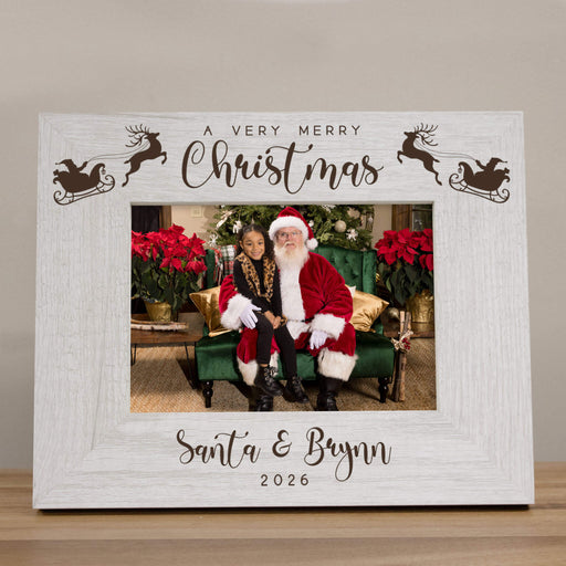 A personalized Christmas photo frame with a distressed white wood-grain finish. The top features the text 'A VERY MERRY Christmas' with reindeer and sleigh silhouettes, and the bottom is personalized with 'Santa & Brynn 2026' in script.