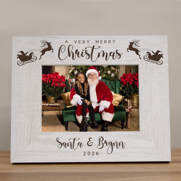 A personalized Christmas photo frame with a distressed white wood-grain finish. The top features the text 'A VERY MERRY Christmas' with reindeer and sleigh silhouettes, and the bottom is personalized with 'Santa & Brynn 2026' in script.