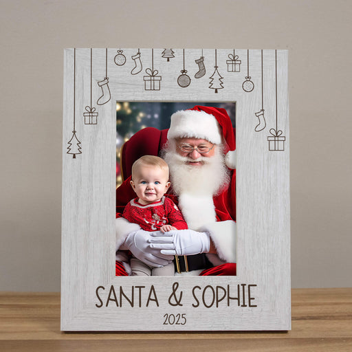 A personalized white wooden picture frame with engraved Christmas-themed hanging icons and the text 'SANTA & SOPHIE 2025' at the bottom, holding a photo of Santa Claus and a baby, displayed on a wooden surface against a neutral beige background.
