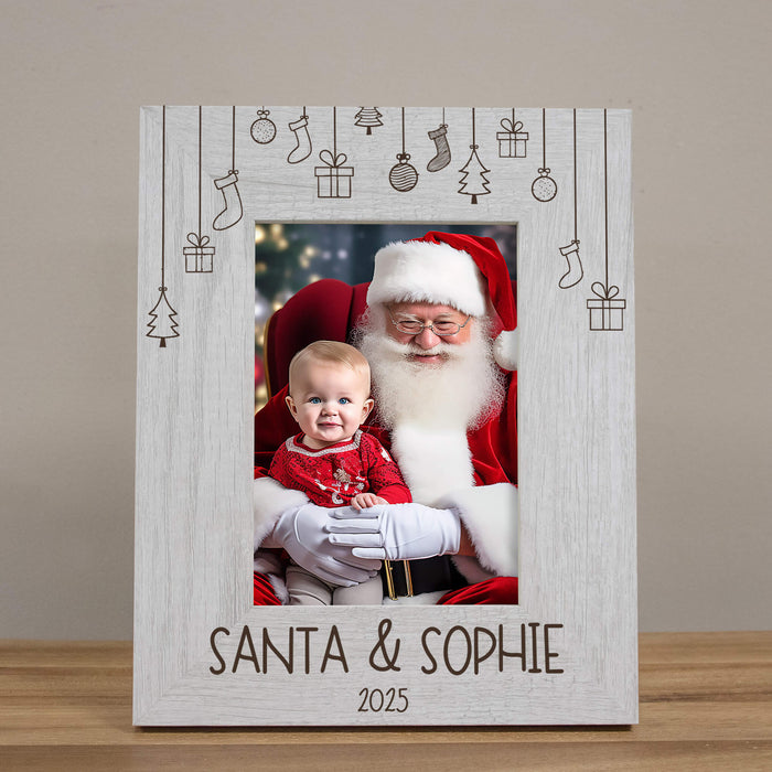 A personalized white wooden picture frame with engraved Christmas-themed hanging icons and the text 'SANTA & SOPHIE 2025' at the bottom, holding a photo of Santa Claus and a baby, displayed on a wooden surface against a neutral beige background.
