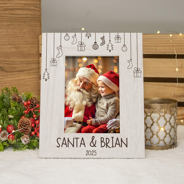 A personalized white wooden Christmas picture frame with engraved line-art icons of stockings, gifts, and trees, featuring a photo of Santa and a child, and the text 'SANTA & BRIAN 2025' at the bottom. The frame is styled on a white surface with holiday greenery and a candle in the background.