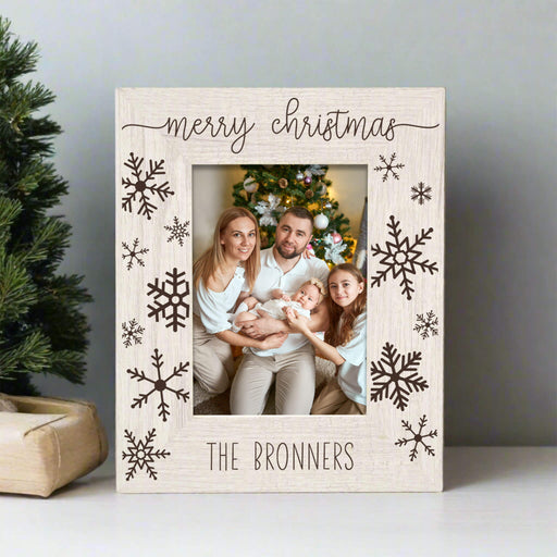 A personalized Christmas photo frame with a light wood grain finish, featuring 'merry christmas' at the top and 'THE BRONNERS' at the bottom, decorated with dark brown snowflakes. A family is visible in the printed photo insert, set against a holiday tree background.