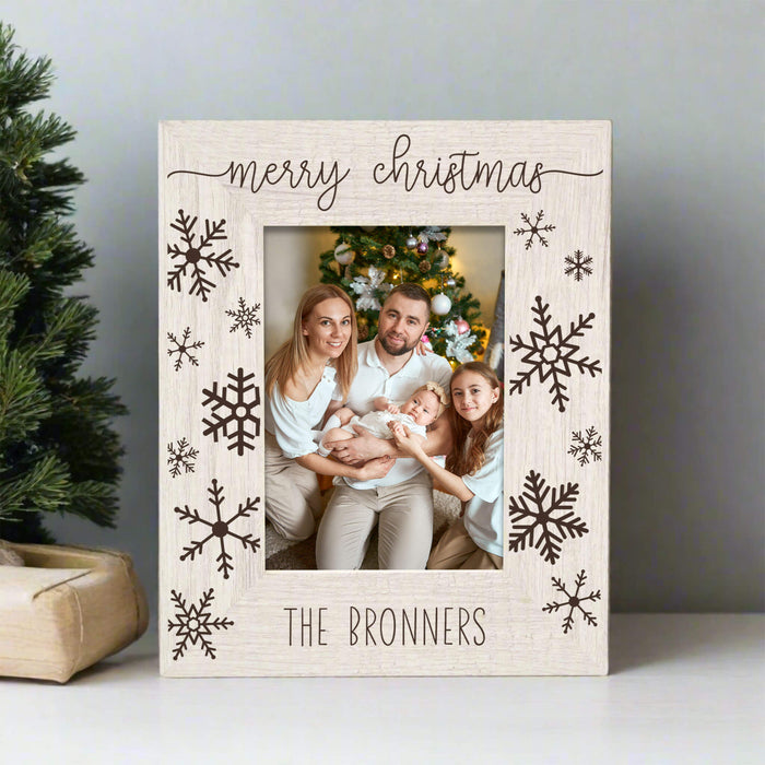 A personalized Christmas photo frame with a light wood grain finish, featuring 'merry christmas' at the top and 'THE BRONNERS' at the bottom, decorated with dark brown snowflakes. A family is visible in the printed photo insert, set against a holiday tree background.