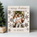 A personalized Christmas photo frame with a light wood grain finish, featuring 'merry christmas' at the top and 'THE BRONNERS' at the bottom, decorated with dark brown snowflakes. A family is visible in the printed photo insert, set against a holiday tree background.