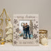 A personalized Christmas picture frame with a light wood finish, featuring 'merry christmas' and 'THE HAWTHORNES' text, decorated with dark brown snowflakes. The frame is styled on a white surface with holiday decorations including a gold star tree, a white ornament, and a candle.
