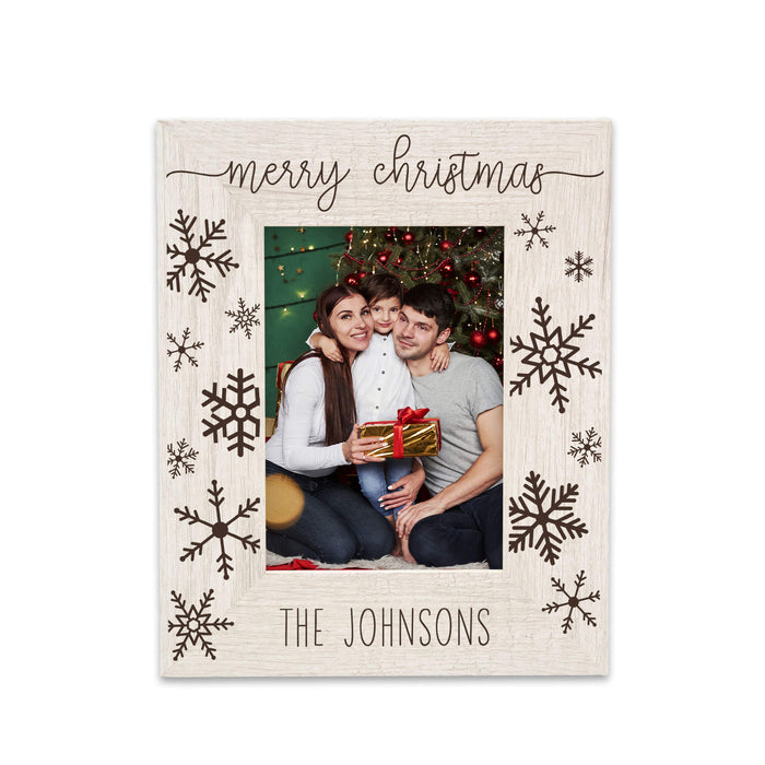 A personalized Christmas photo frame with a white wood-grain border, featuring dark brown snowflakes, the text 'merry christmas' at the top, and 'THE JOHNSONS' at the bottom. A custom photo of a family is displayed in the center.