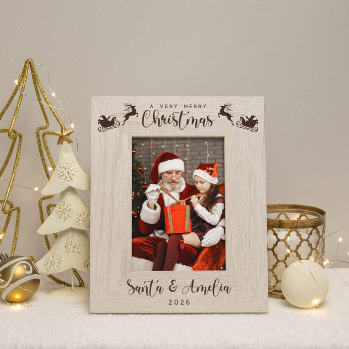 A personalized wooden Christmas photo frame with a light natural wood finish, featuring engraved holiday illustrations and the text 'A VERY MERRY Christmas Santa & Amelia 2026'. The frame is displayed on a white surface with festive holiday decorations, including a gold star, a white decorative tree, and a gold candle holder.