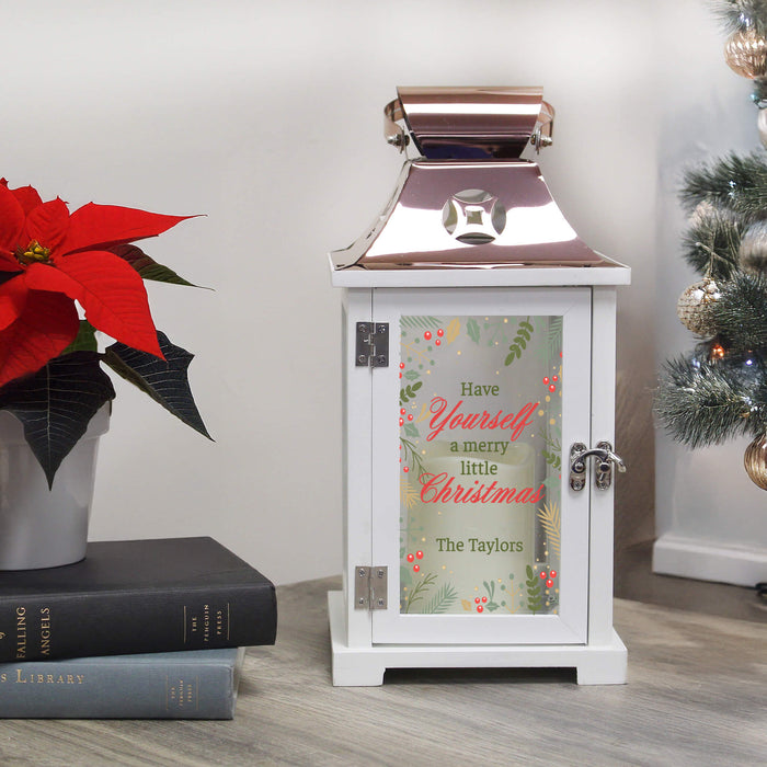A white decorative lantern with a copper metallic roof and a personalized glass door featuring a Christmas message, styled on a wooden floor next to a poinsettia and a Christmas tree.