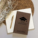 Brown notebook with graduation design and text on a white surface with a pen and dried flowers.