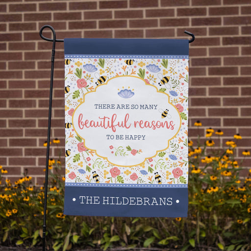 Decorative garden flag with floral bee design and text in front of a brick wall.