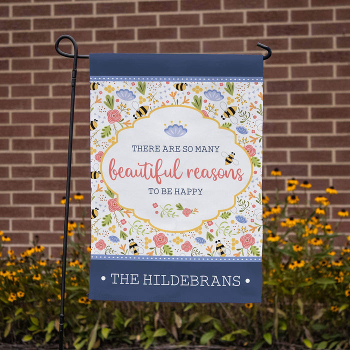 Decorative garden flag with floral bee design and text in front of a brick wall.