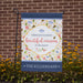 Decorative garden flag with floral bee design and text in front of a brick wall.