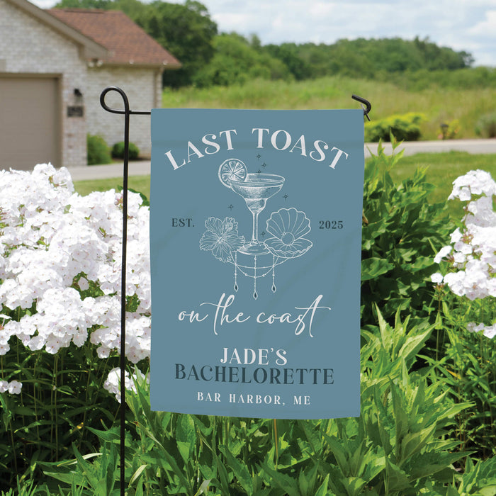 Garden flag with 'Last Toast on the Coast' text and design, set against a natural background with flowers and a house.