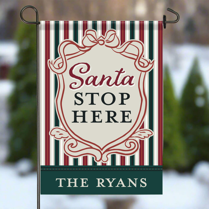 A vertical garden flag with a holiday design, featuring red, green, and white stripes, a central cream shield with the text 'Santa STOP HERE' in red and dark green, and a dark green bottom banner reading 'THE RYANS'. The flag is displayed on a metal stand against a blurred winter outdoor background.