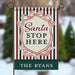 A vertical garden flag with a holiday design, featuring red, green, and white stripes, a central cream shield with the text 'Santa STOP HERE' in red and dark green, and a dark green bottom banner reading 'THE RYANS'. The flag is displayed on a metal stand against a blurred winter outdoor background.