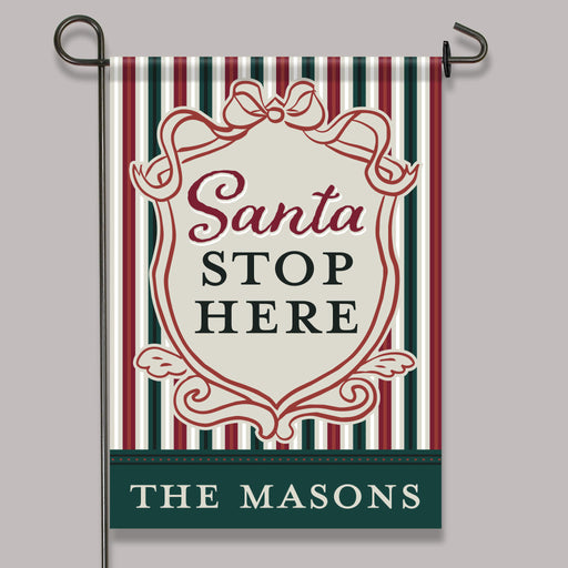 A vertical garden flag hanging from a black metal stand against a plain light gray background. The flag has a holiday-themed design with red, dark green, and cream vertical stripes, a central cream shield with the words 'Santa STOP HERE' in red and dark green, and a dark green bottom banner reading 'THE MASON'S' in white.