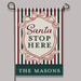 A vertical garden flag hanging from a black metal stand against a plain light gray background. The flag has a holiday-themed design with red, dark green, and cream vertical stripes, a central cream shield with the words 'Santa STOP HERE' in red and dark green, and a dark green bottom banner reading 'THE MASON'S' in white.