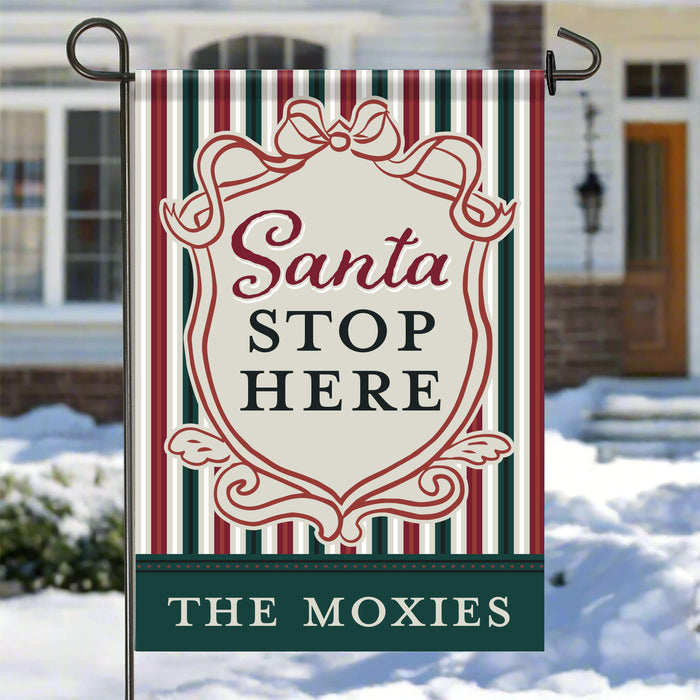 A vertical garden flag hanging from a metal stand in a snowy outdoor setting. The flag has a holiday design with red, green, and cream vertical stripes, a central cream shield with the words 'Santa STOP HERE' in red and black, and a dark green bottom banner reading 'THE MOXIES' in white.