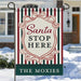 A vertical garden flag hanging from a metal stand in a snowy outdoor setting. The flag has a holiday design with red, green, and cream vertical stripes, a central cream shield with the words 'Santa STOP HERE' in red and black, and a dark green bottom banner reading 'THE MOXIES' in white.