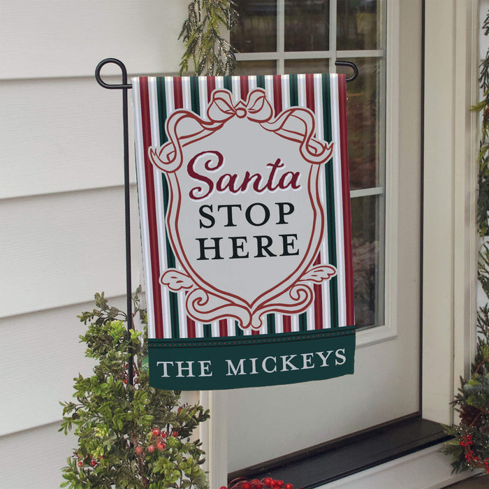 A decorative holiday garden flag hanging from a black metal stand on a house door. The flag has red, green, and white stripes, an ornate white crest with red script reading 'Santa STOP HERE', and a dark green banner at the bottom reading 'THE MICKEYS'.