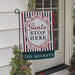 A decorative holiday garden flag hanging from a black metal stand on a house door. The flag has red, green, and white stripes, an ornate white crest with red script reading 'Santa STOP HERE', and a dark green banner at the bottom reading 'THE MICKEYS'.