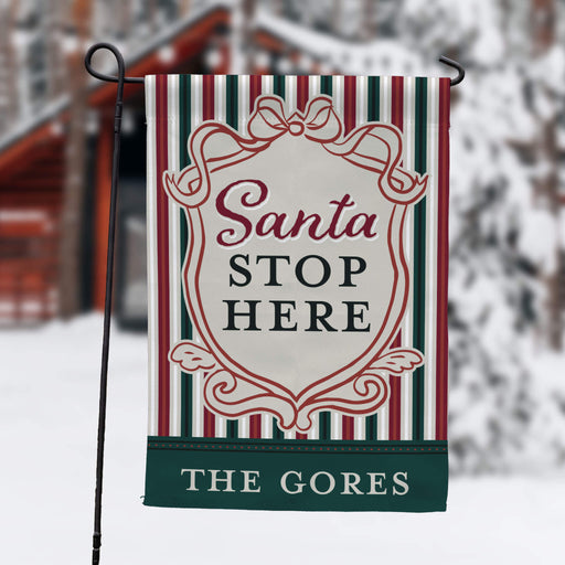 A vertical garden flag on a black metal stand, featuring a holiday design with red, green, and white stripes, a cream central shield with the text 'Santa STOP HERE', and a dark green bottom banner reading 'THE GORES'. The background is a blurred snowy outdoor scene.