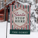 A vertical garden flag on a black metal stand, featuring a holiday design with red, green, and white stripes, a cream central shield with the text 'Santa STOP HERE', and a dark green bottom banner reading 'THE GORES'. The background is a blurred snowy outdoor scene.
