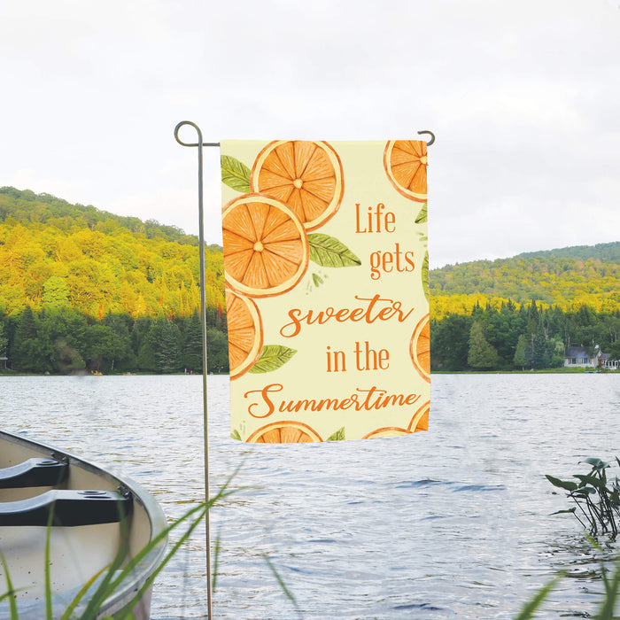 Flag with orange slices and 'Life gets sweeter in the Summertime' text in a scenic lake setting.