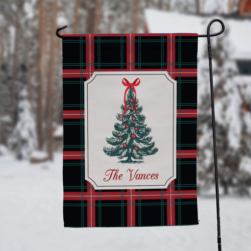 A personalized holiday garden flag hanging from a black metal stand in a snowy outdoor setting. The flag features a black and red plaid pattern with a central white label containing a green Christmas tree illustration and the text 'The Vances'.
